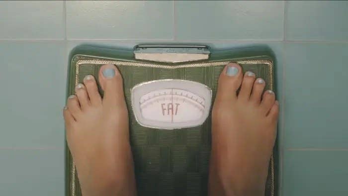 Feet standing on a bathroom scale that has no numbers, it just reads "fat"