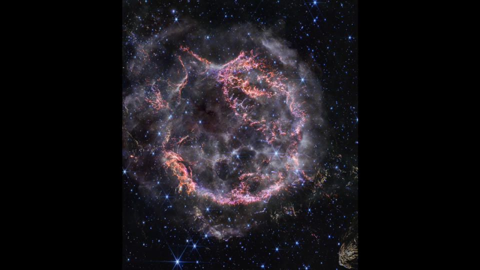 Cassiopeia A, a circular-shaped cloud of gas and dust with complex structure. The inner shell is made of bright pink and orange filaments studded with clumps and knots that look like tiny pieces of shattered glass. Around the exterior of the inner shell, particularly at the upper right, there are curtains of wispy gas that look like campfire smoke. The white smoke-like material also appears to fill the cavity of the inner shell, featuring structures shaped like large bubbles. Around and within the nebula, there are various stars seen as points of blue and white light. Outside the nebula, there are also clumps of yellow dust, with a particularly large clump at the bottom right corner that appears to have very detailed striations.