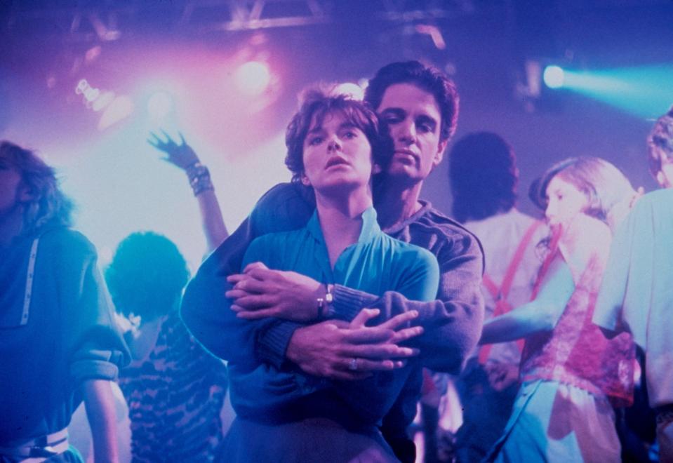 A couple embraces closely while dancing in a crowded nightclub. Bright lights and energetic dancers surround them