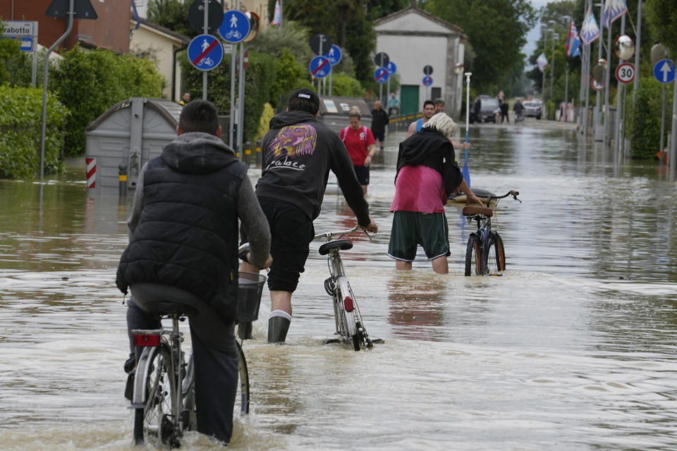 People pedal in a flooded street in Barbiano di Cotignola, Italy, Thursday, May 18, 2023. Exceptional rains Wednesday in a drought-struck region of northern Italy swelled rivers over their banks, killing at least nine people. Rescue crews worked Thursday to reach isolated towns and villages in northern Italy that were cut off from highways, electricity and cellphone service following heavy rains and flooding, as farmers warned of "incalculable" losses and authorities began mapping out cleanup and reconstruction plans. (AP Photo/Luca Bruno)