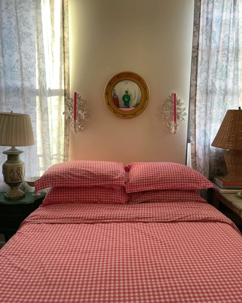 Mirror and candle sconces above bed with red gingham bedding flanked by lamps and windows in white bedroom.