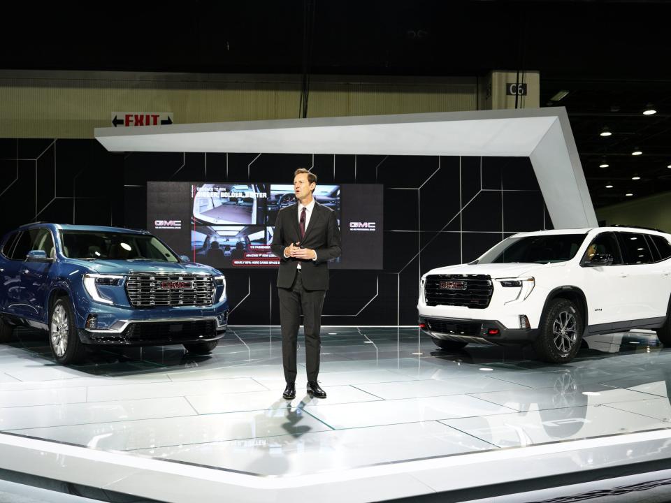 A blue SUV and a white SUV are positioned on a shiny auto show floor. A man speaks in between the two.