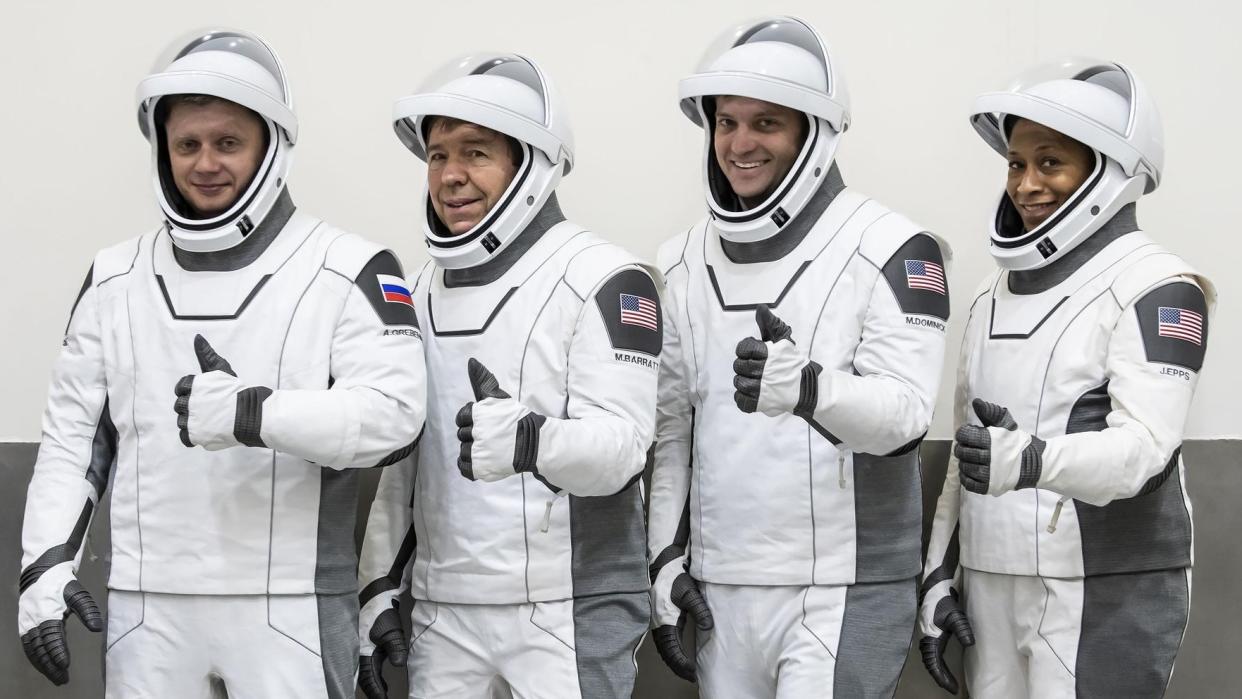  Four astronauts with white spacesuits and helmets giving a thumbs up while standing in a row. 