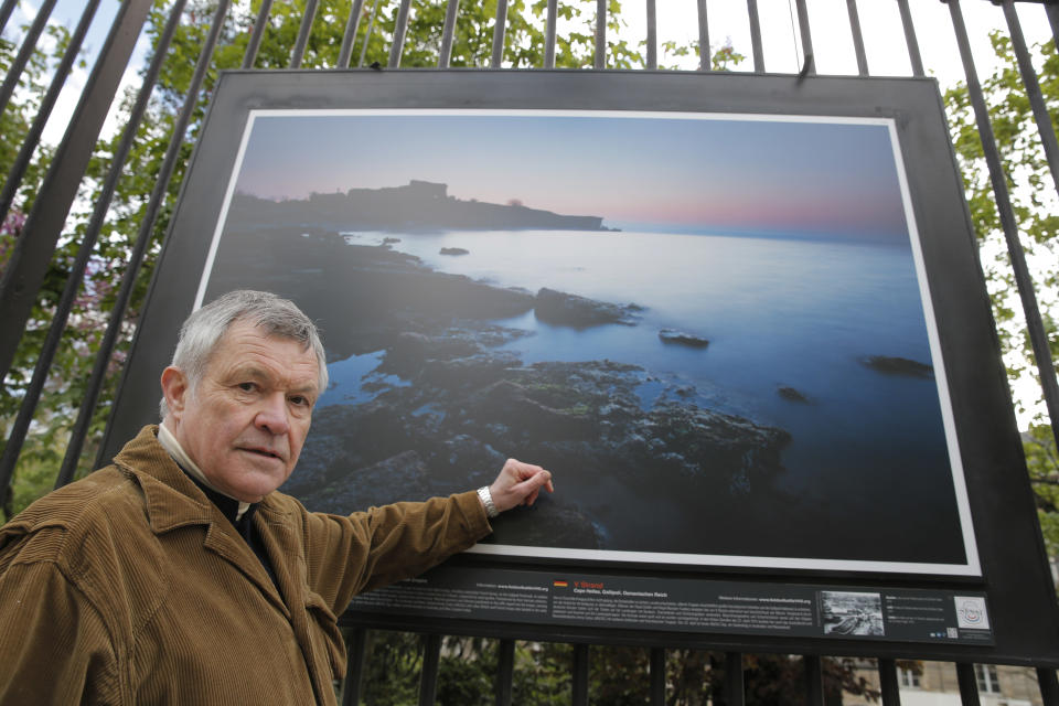 British photographer Michael St Maur Sheil poses in front of one of his pictures displayed at the Paris Luxembourg gardens, Tuesday, April 8, 2014, as part of an exhibition " Fields of Battle - Lands of Peace 14-18 ". The picture displayed is a modern take at the location of the Cape Helles landings on the Gallipoli peninsula. Captured over a period of seven years, Michael’s photography combines a passion for history and landscape and presents a unique reflection on the transformation of the battlefields of the Great War into the landscape of modern Europe. (AP Photo/Christophe Ena)