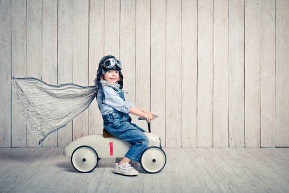 A small boy rides a toy car with a cape on