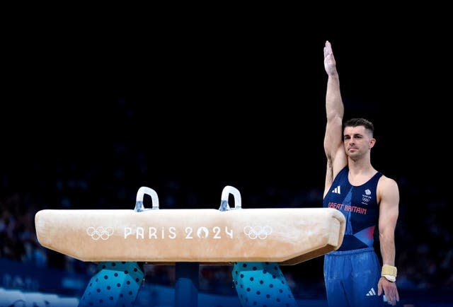 Max Whitlock with his right arm raised upright as he stands behind a pommel horse