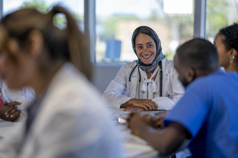 Medical provider wearing a hijab smiling at other medical providers sitting at a table wearing scrubs and white coats.