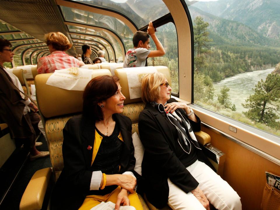 Guests sit on a luxury train with panoramic views of mountains out wide windows on the right.