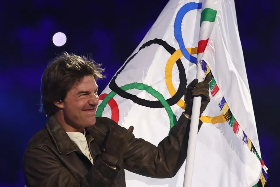 Tom Cruise holding an Olympic flag with a smile. He is wearing a brown leather jacket and gloves