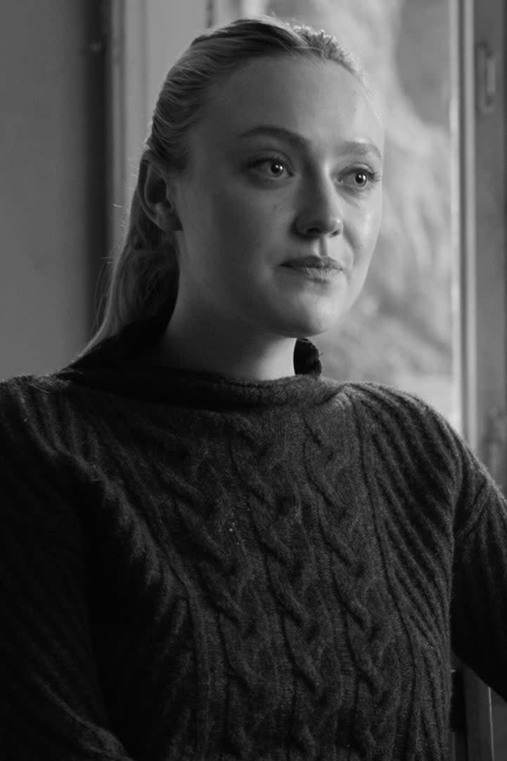 Dakota Fanning sits by a table in a room with a window, wearing a cable-knit sweater. The table is cluttered with papers and books