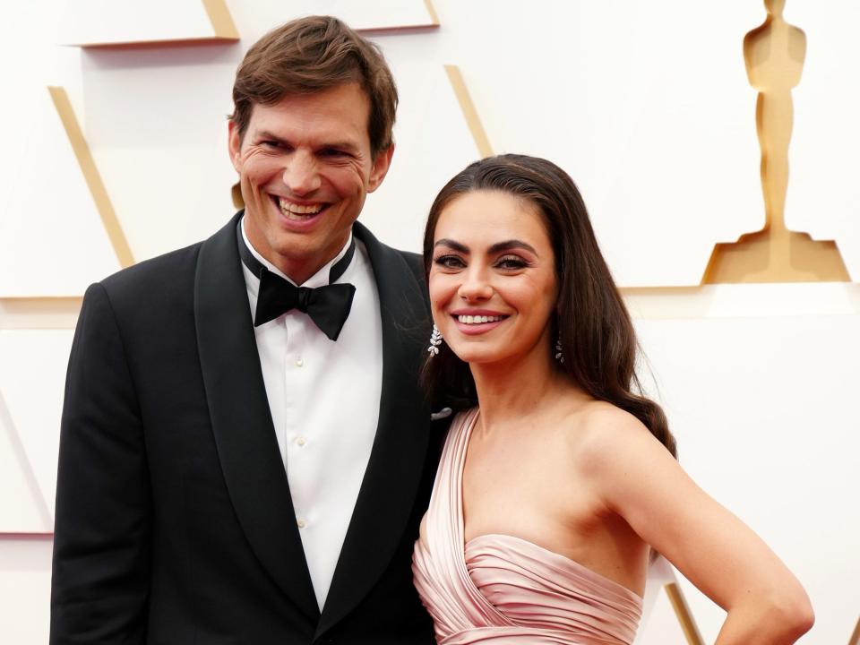 Ashton Kutcher, in a tuxedo, and Mila Kunis, in a strapless blush gown, smile while posing on the red carpet together.