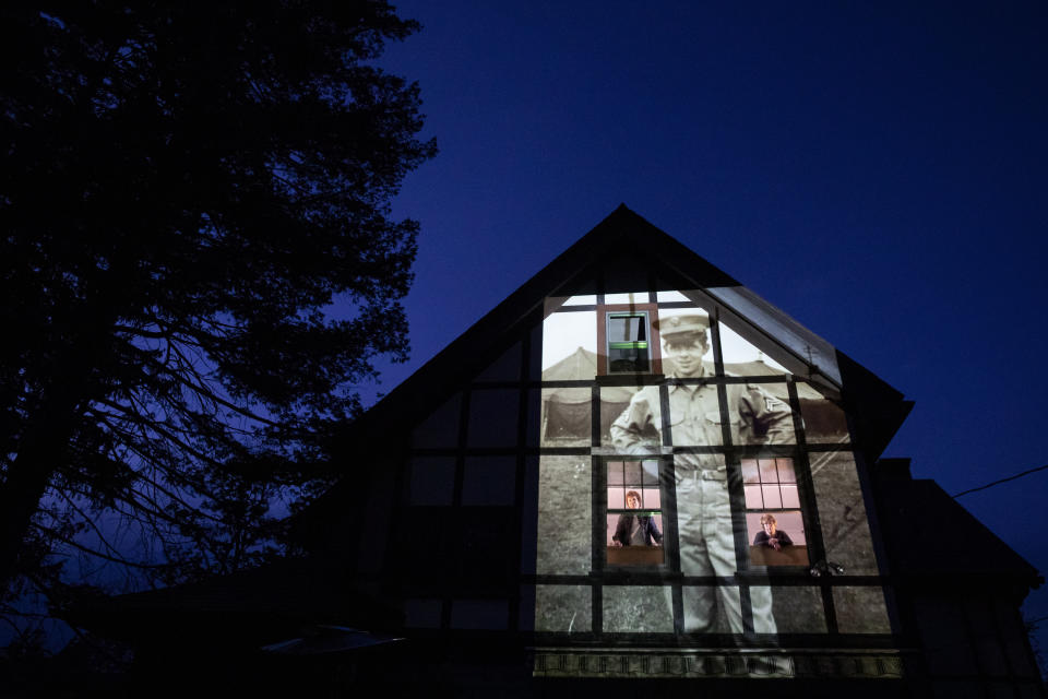 An image of veteran Alfred Healy is projected onto the home of his daughter, Eileen Driscoll, left, as she looks out the window with her sister, Patricia Creran, in Holyoke, Mass., Thursday, May 7, 2020. Healy, a U.S. Army Korean War veteran and resident of the Soldier's Home in Holyoke, Mass., died from the COVID-19 virus at the age of 91. Seeking to capture moments of private mourning at a time of global isolation, the photographer used a projector to cast large images of veterans on to the homes as their loved ones are struggling to honor them during a lockdown that has sidelined many funeral traditions. (AP Photo/David Goldman)