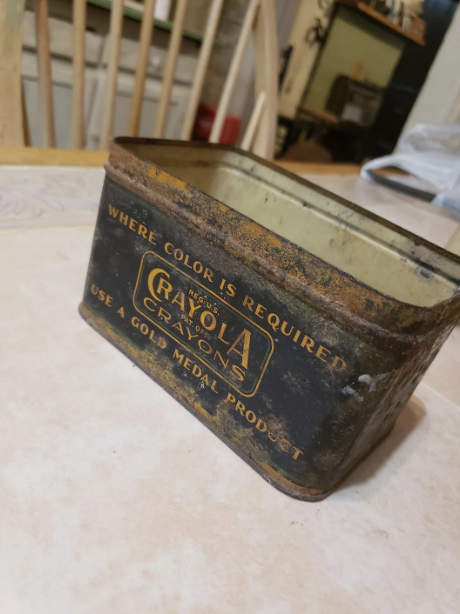 An empty, weathered Crayola Crayons metal container is lying on its side on a white table. The text on the container reads, "WHERE COLOR IS REQUIRED" and "USE A GOLD MEDAL PRODUCT."