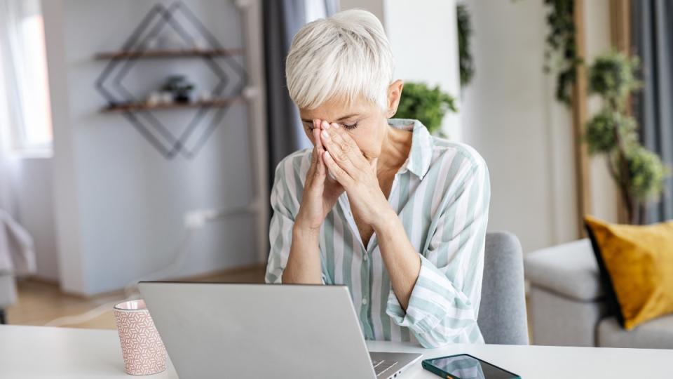 A grey haired woman with her hands on her head in front of a laptop who is experiencing afternoon slump