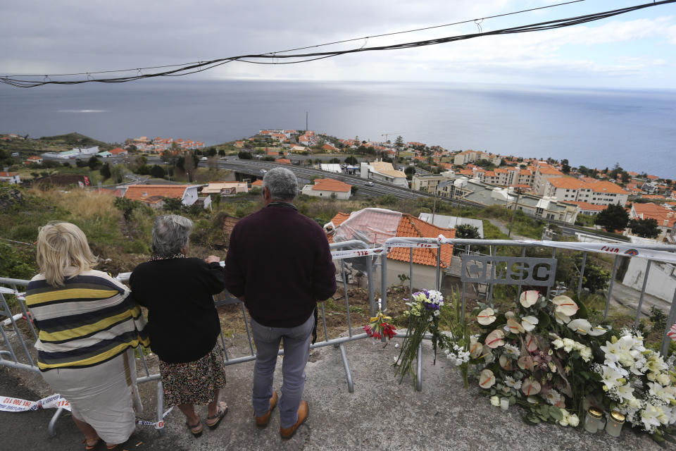 People stand by flowers placed at the spot where a tourist bus veered off the road crashing into a house, seen below, killing 29 people in Funchal, the capital of Portugal's Madeira Island, Friday April 19, 2019. A German plane is expected to arrive Friday in Madeira to take home survivors from the bus accident that killed 29 tourists after it veered off the road and plunged down a slope. All the deceased were German. The bus carried 55 people, including a Portuguese driver and guide. Sixteen people remain hospitalized, but authorities said all of them are out of danger. (AP Photo/Armando Franca)