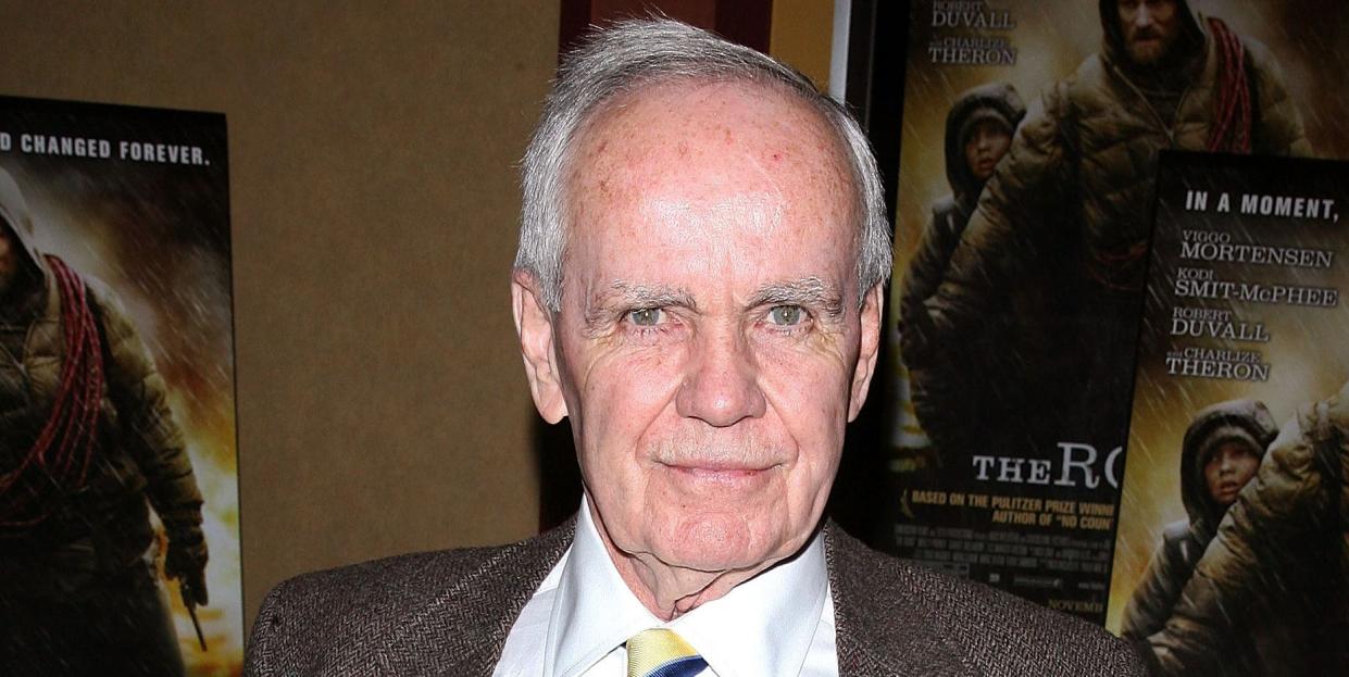 cormac mccarthy, wearing a brown suit and blue and yellow striped tie, looking directly into the camera while standing in front of three posters for the movie the road