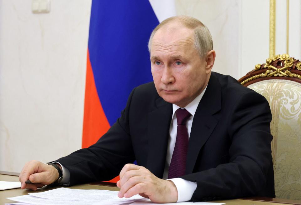 President Vladimir Putin sitting in a gilded chair with a Russian flag behind him, looking glum.