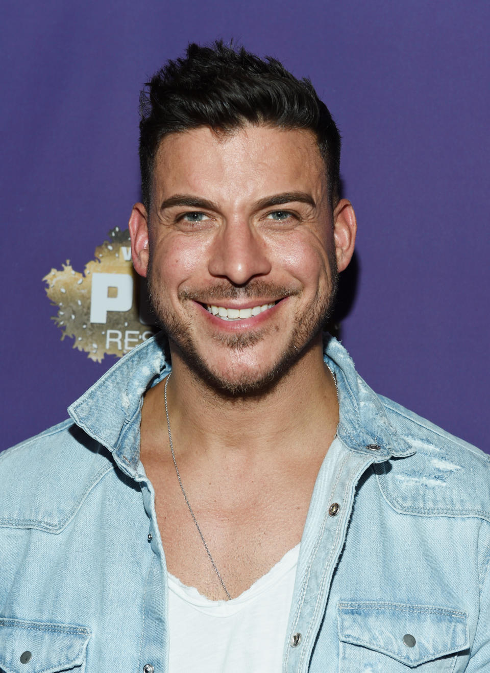 Jax Taylor smiles wearing a denim jacket over a white shirt at the event against a purple backdrop.