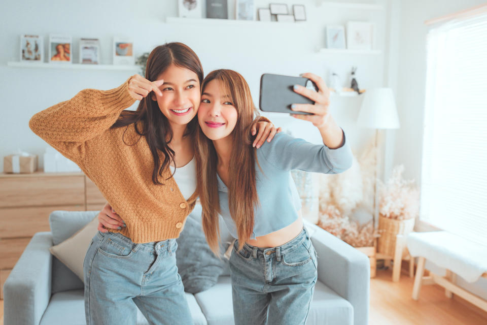 Two happy asian women blogger shooting social media stories or video calling in mobile app and enjoying rest on comfortable sofa at home. selfie