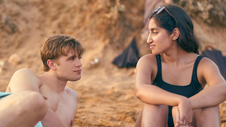 A man and woman in bathing suits look at each other while sitting on a beach; still from "One Day"