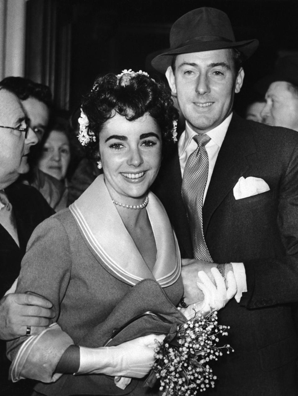 Elizabeth Taylor, holding a bouquet with white gloves, smiles as she holds hands with smiling husband Michael Wilding.