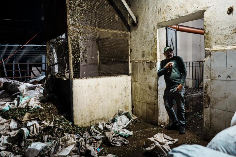 A man with a cup leaning against a doorway in a room strewn with bags and olive debris