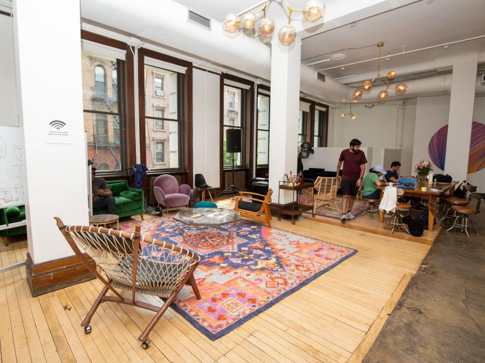 A room with large windows, a colorful rug, couches, chairs. People are working at a long table nearby.