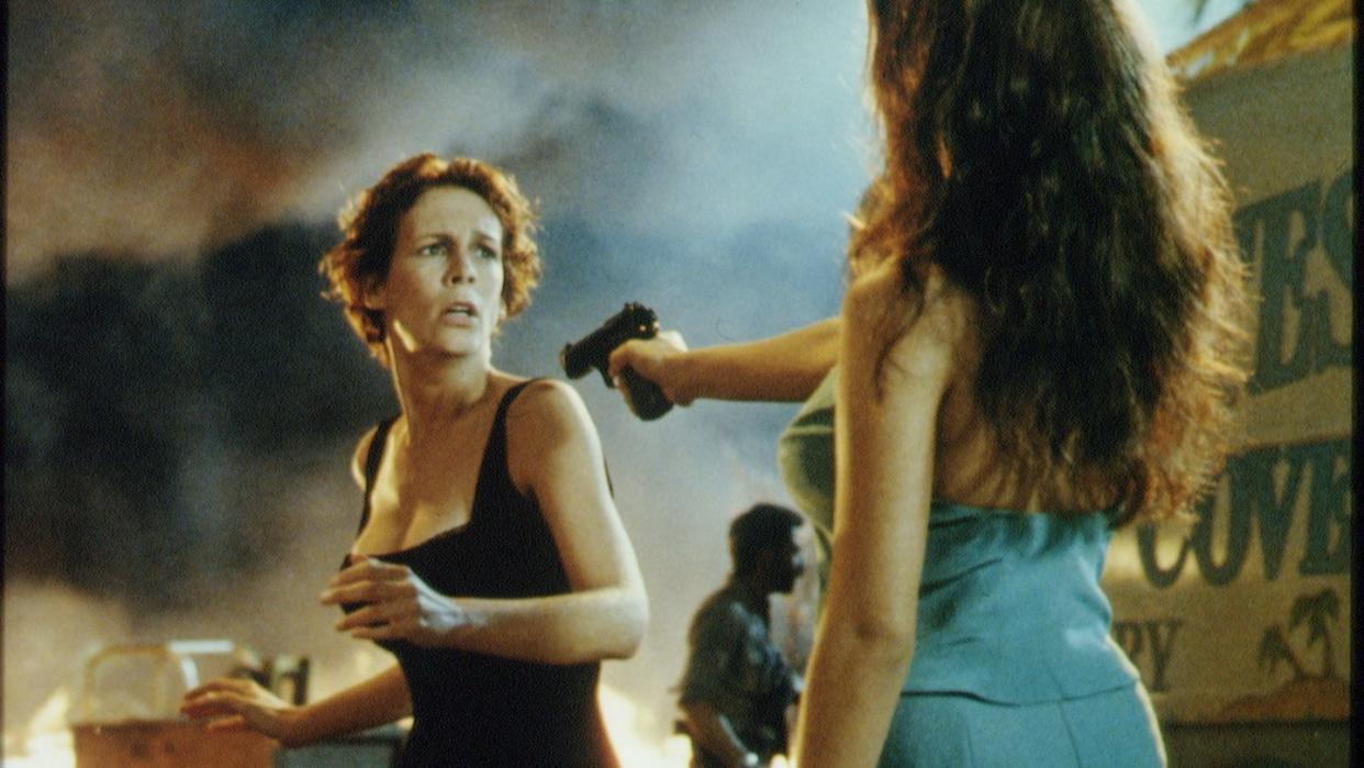 a publicity photo from the movie true lies, featuring jamie lee curtis reacting with concern as a woman points a gun at her