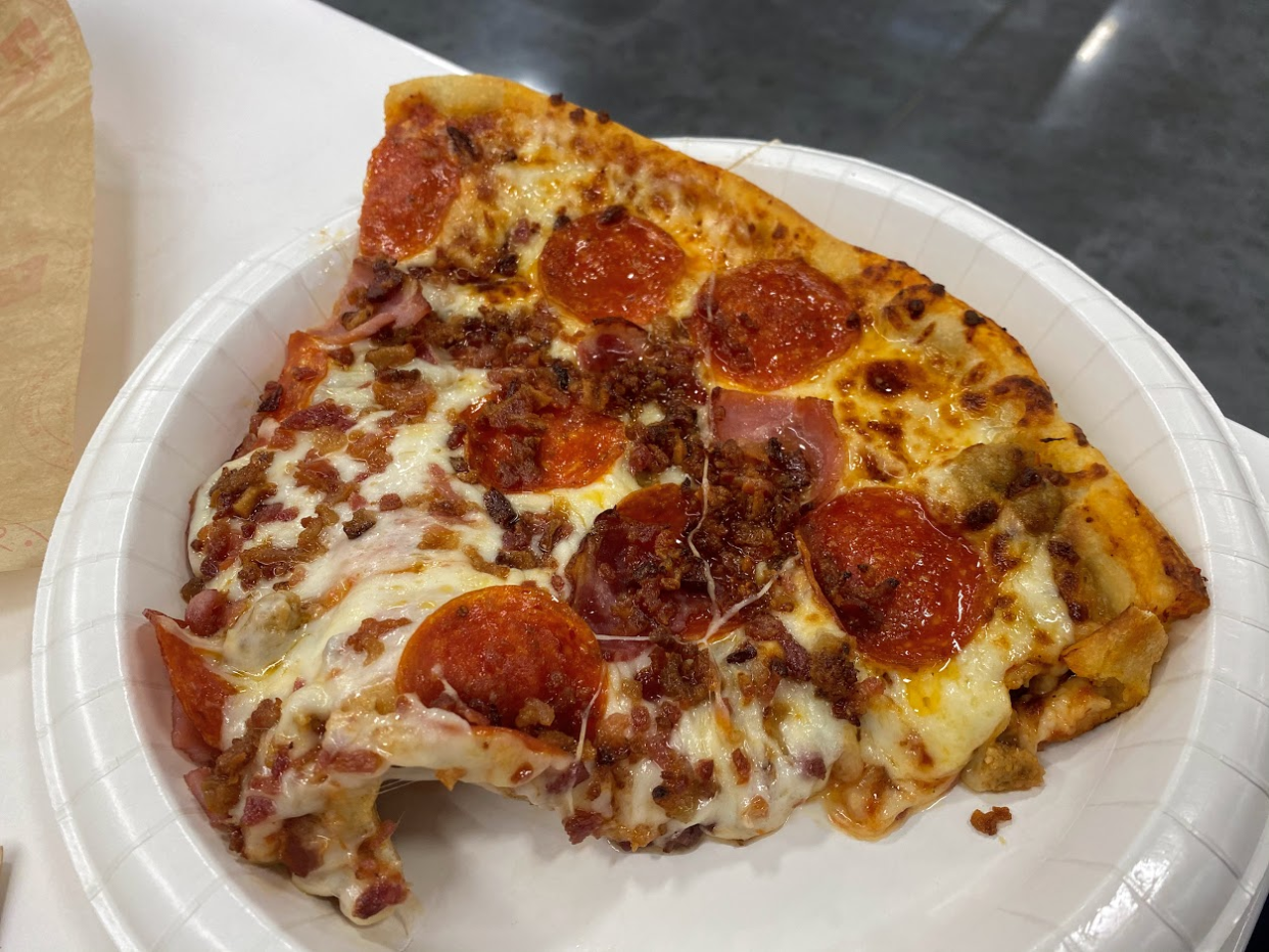 Slice of a pepperoni and sausage pizza on a white paper plate at the Sam's Club cafe, on a white table with a dark grey floor on the top right background