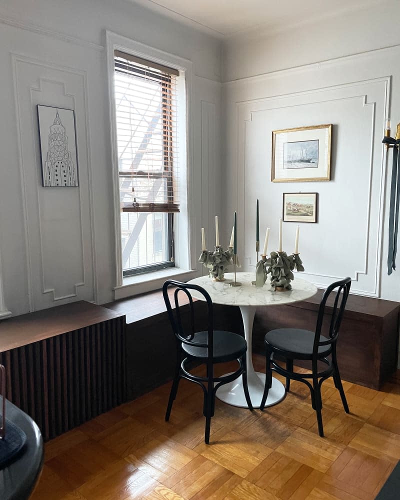 There is a marble-top tulip table and wooden chairs next to a wooden window bench in the corner of the room, and art is hung on the walls with decorative moldings.