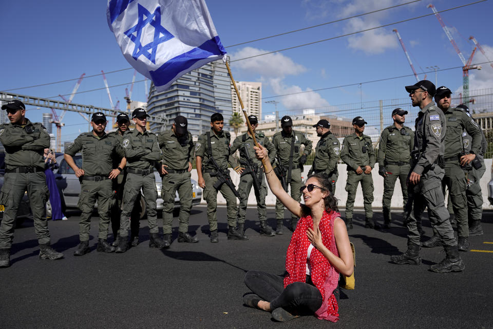 A demonstrator waves the Israeli flag seated on a highway while flanked by paramilitary border police during a protest against plans by Prime Minister Benjamin Netanyahu's government to overhaul the judicial system, in Tel Aviv, Israel, Thursday, March 9, 2023. (AP Photo/Ariel Schalit)