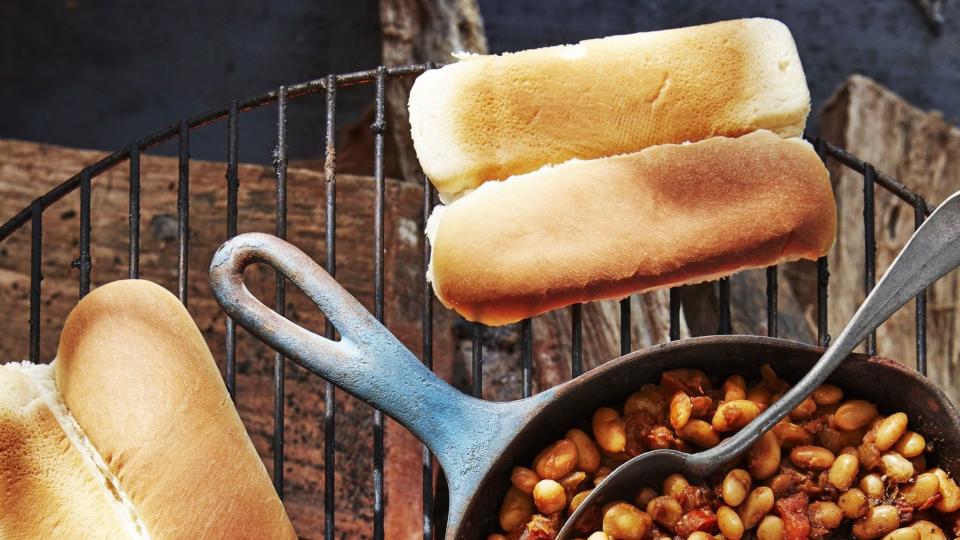 hot dogs and buns on a grill with a cast iron pan full of beans with a spoon