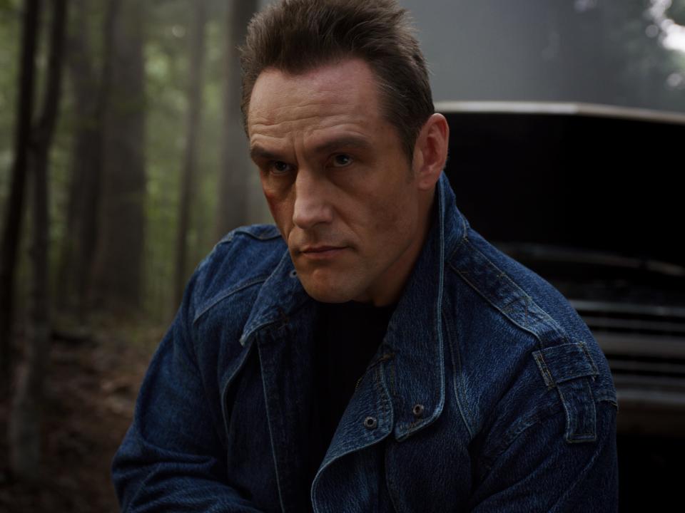 grigori in stranger things, standing in front of a car with its hood up in the forest