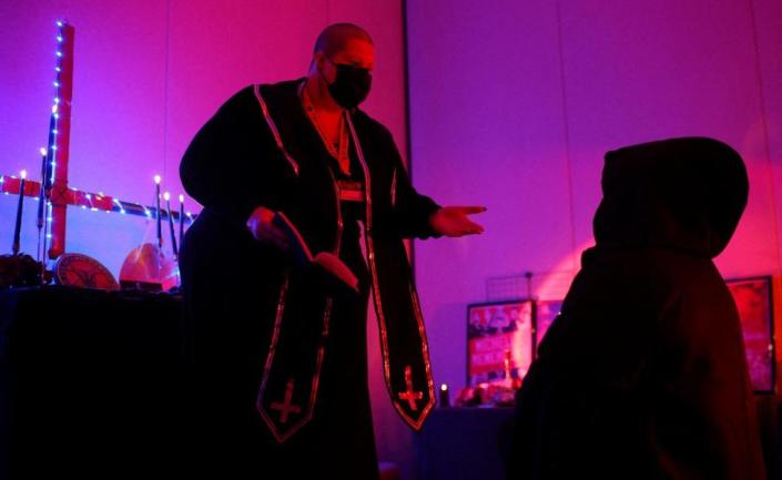 Minister Rose d&#39;Arc performs an Unbaptism Ceremony in a black robe edged with upside down crosses. An altar decked in blue-white lights also with an upside down cross on it stands behind. The subject of the ceremony kneels down in a black robe and hood.