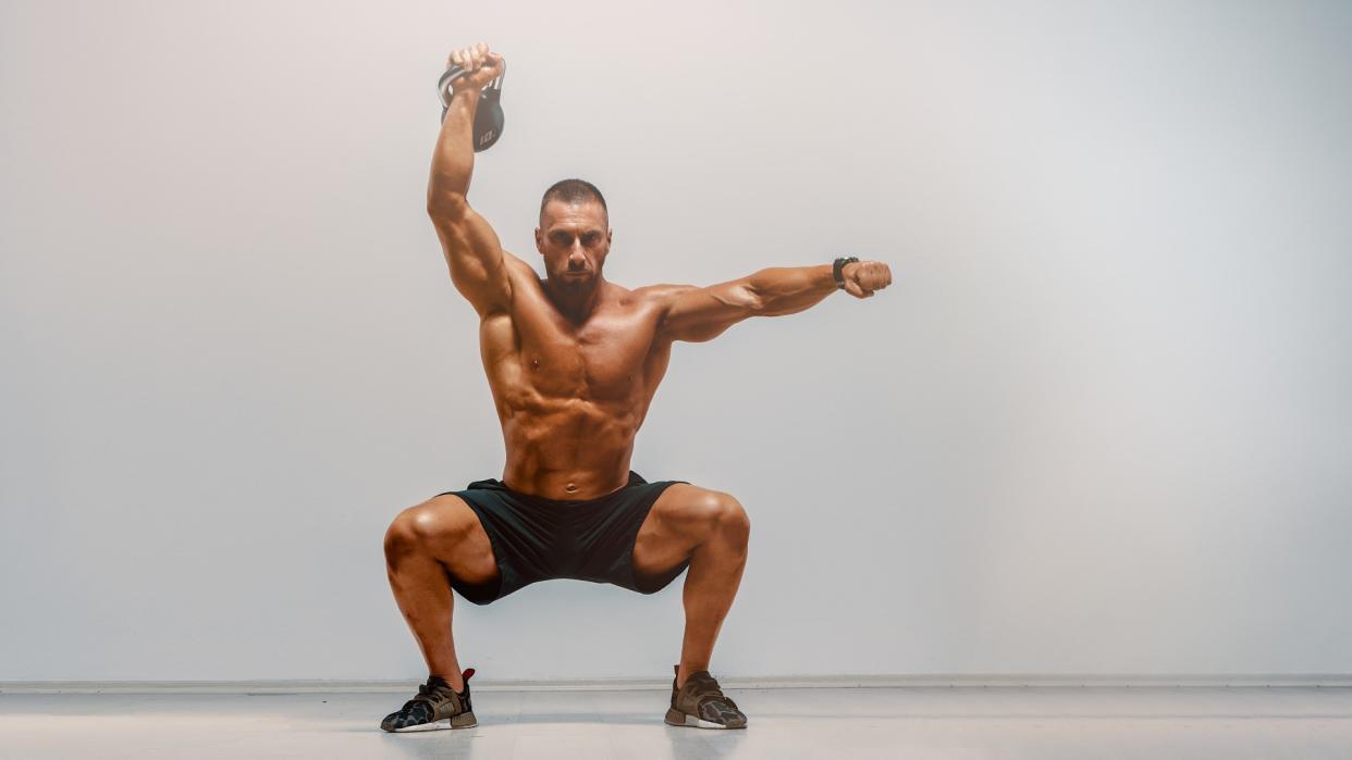  Man against grey backdrop performing a single arm overhead kettlebell squat with right arm raised. 