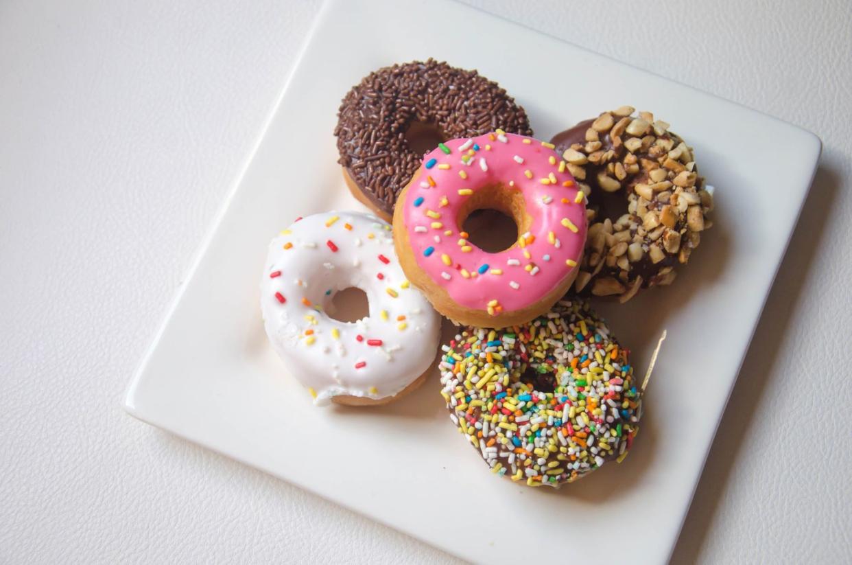 five doughnuts on a square white plate with multi colored icing and nuts or spinkles