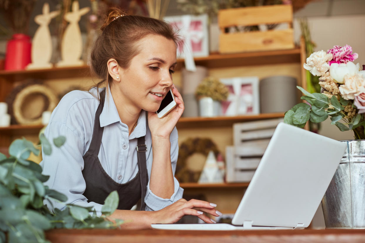 lifetime ISA Portrait of florist in apron working at her own flower shop, using laptop and calling on the smart phone. She is leaning on wooden counter. She is arranging logistics, and delivery, taking orders