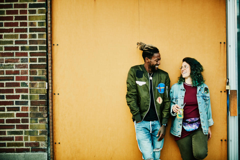 Two people leaning against a wall, smiling and engaged in a conversation, dressed in casual jackets and jeans