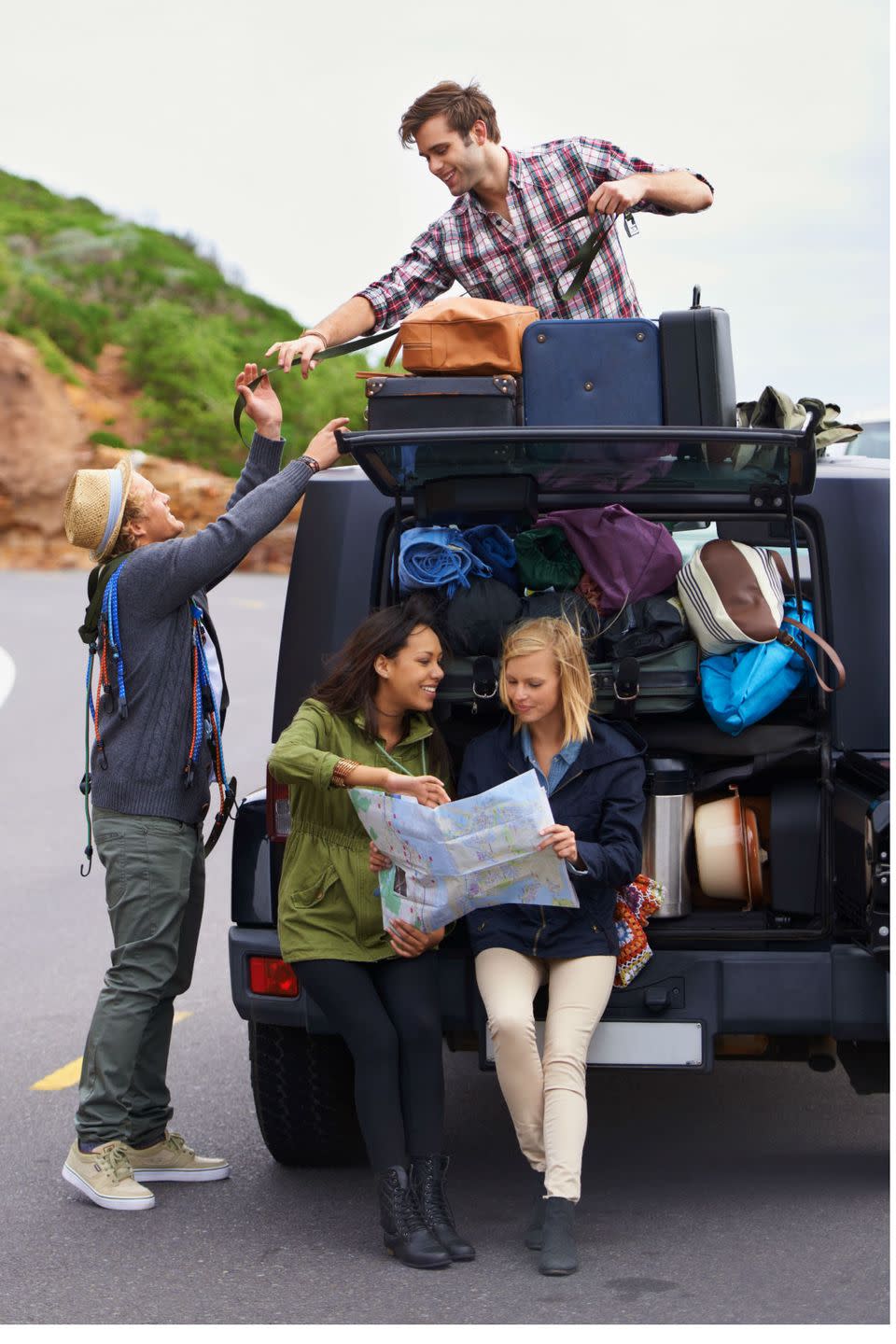 two young men strap luggage to the top of a car while two young women sit on the bumper reviewing a map for a road trip, a summer bucket list idea