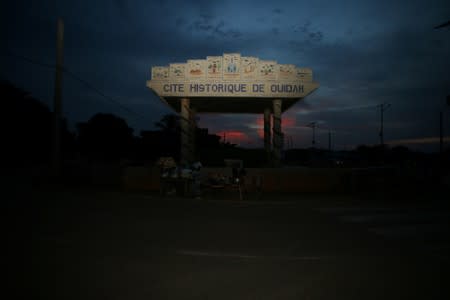 Woman sells food on the street infront of a monument with an inscrption Ouidah Historic City along Ouidah-Togo highway