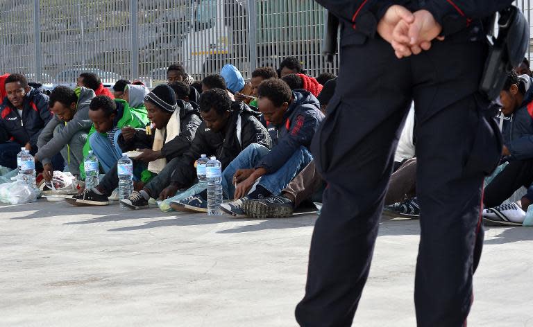 An Italian policeman stands guard as migrants eat while waiting at the port of Lampedusa to board a ferry bound for Porto Empedocle in Sicily on February 20, 2015