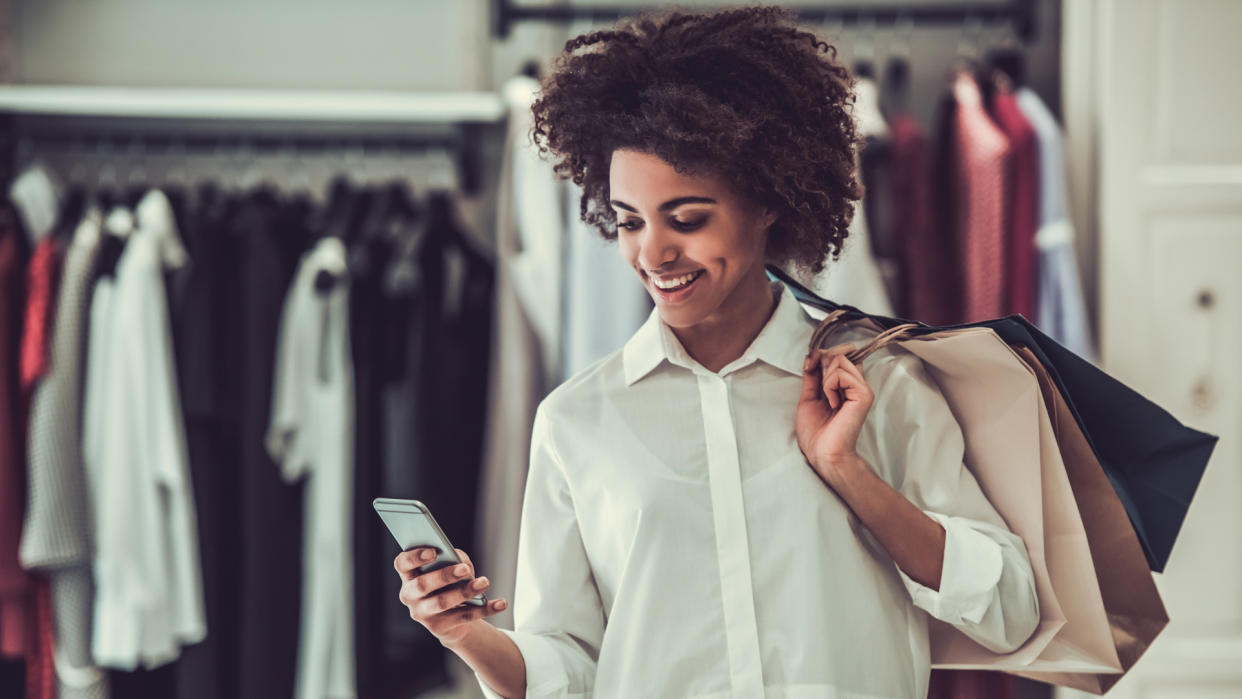 Girl is holding shopping bags, using a smart phone and smiling while doing shopping in female boutique.