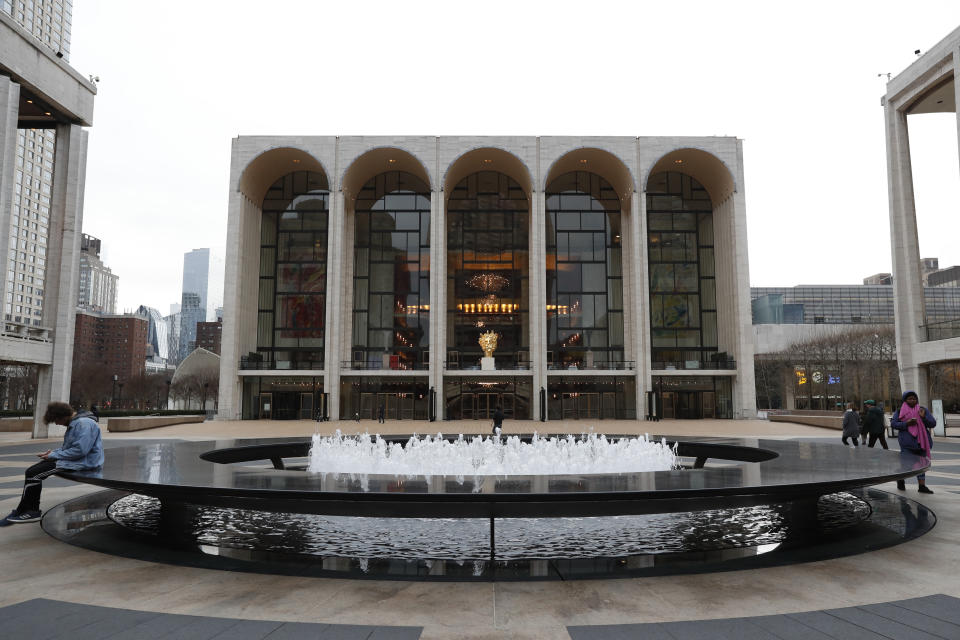 FILE - This March 12, 2020 file photo shows a few people at Josie Robertson Plaza in front of The Metropolitan Opera house, background center, at Lincoln Center in New York. The Metropolitan Opera is canceling the rest of its season and stopping pay of the orchestra, chorus and other unionized employees at the end of March due to the new coronavirus. Met general manager Peter Gelb said he is giving up his $1.45 million salary until normal operations resume and higher-paid members of his administrative staff are having their pay reduced, by 10-50% for everyone earning more than $125,000. (AP Photo/Kathy Willens, File)