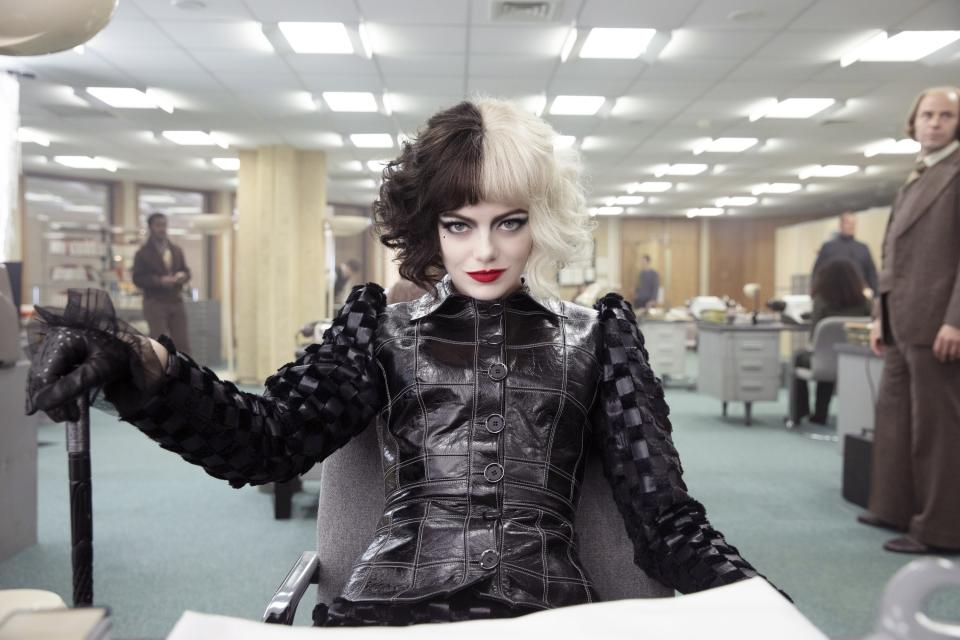 Emma Stone dressed as Cruella de Vil in a stylish, black leather outfit with puffed sleeves, sits confidently in an office setting