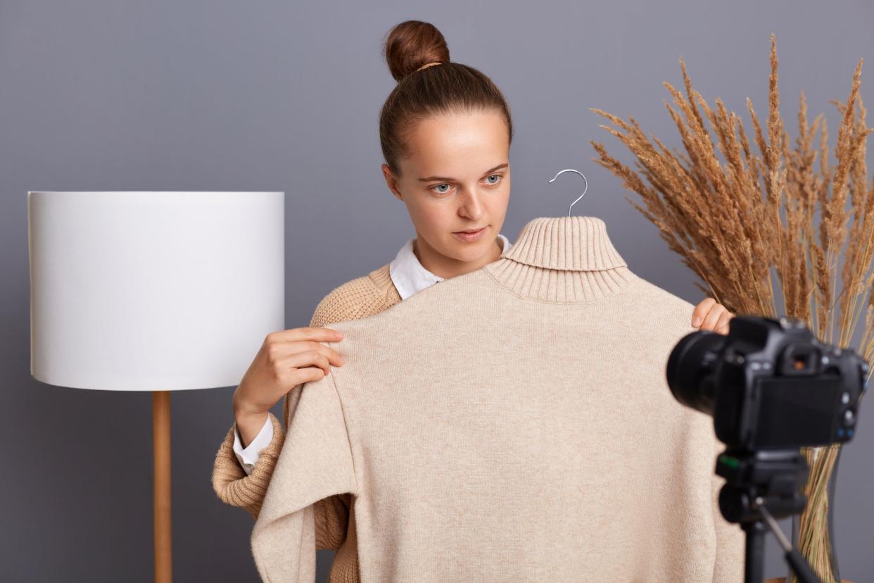 Image of concentrated serious confident woman stylist with bun hairstyle recording video, using camera on tripod, shooting new content for her fashion vlog, showing new jumper, selling clothe online.