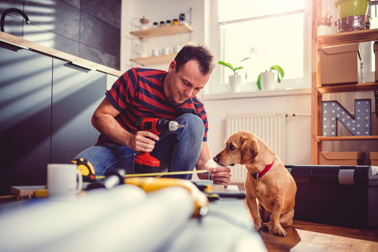 Man holding a cordless drill and measuring cabinets on the floor to install, in the kitchen with a small brown dog, light coming into through the window