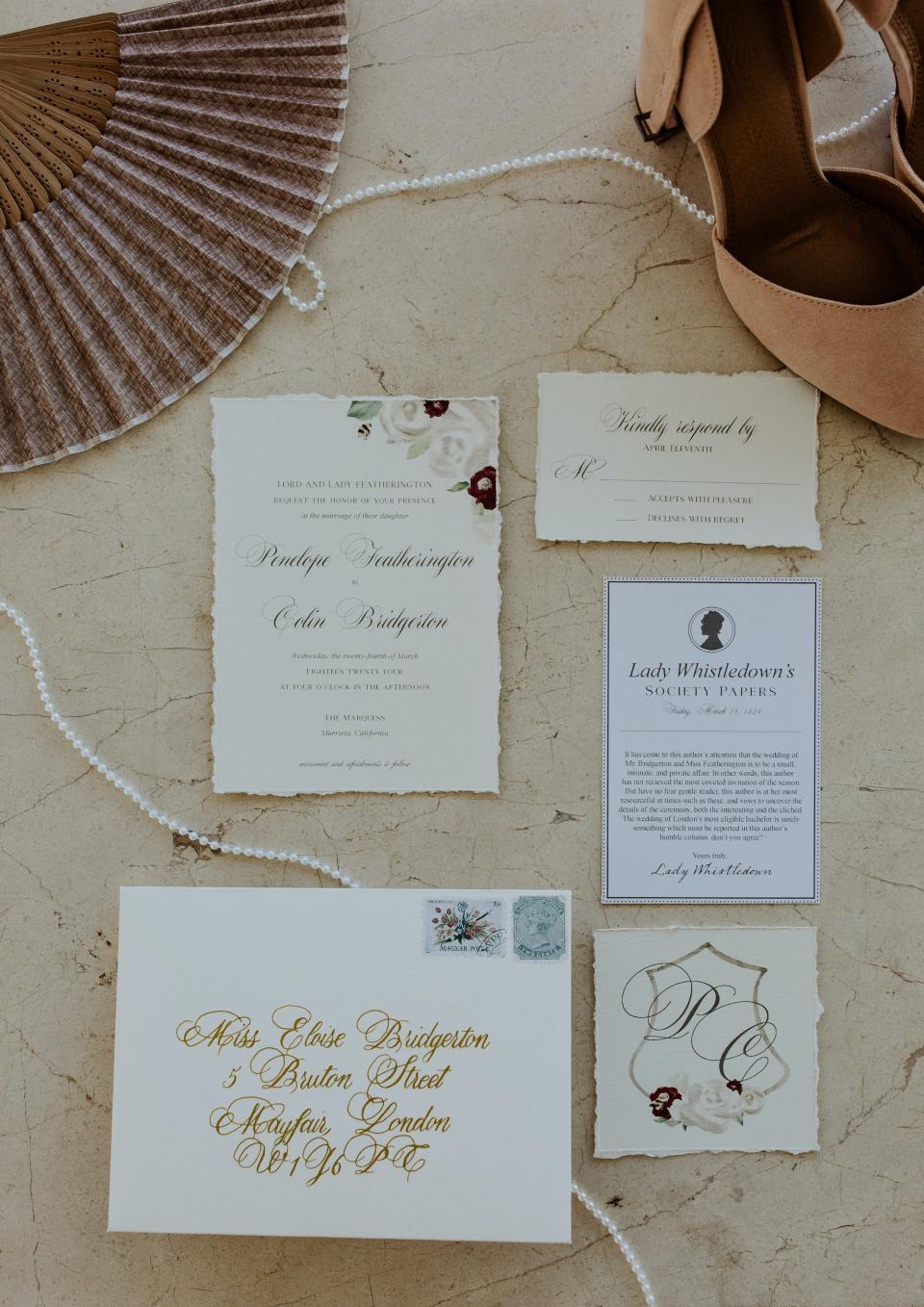 Wedding invitations sit on a table next to a fan, a string of pearls, and a tan shoe.