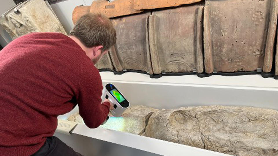 A man in a red shirt holds a scanner with a green light. He is scanning the concrete-like remains of a human burial that sits in a white container against a wall.