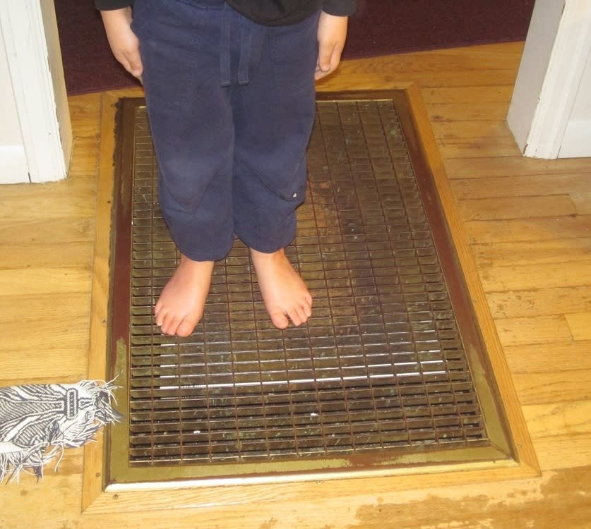 A child in blue pants stands barefoot on a floor vent in a home