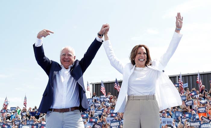 Kamala Harris and Tim Walz stand holding hands, celebrating in front of a crowd with American flags and "We Win" signs